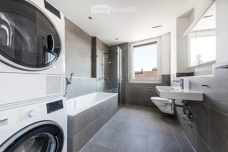 Modern bathroom with a bathtub, toilet, sink, and washing machine. Features a large window with a view of a garden, sleek fixtures, and a glass-enclosed shower area.