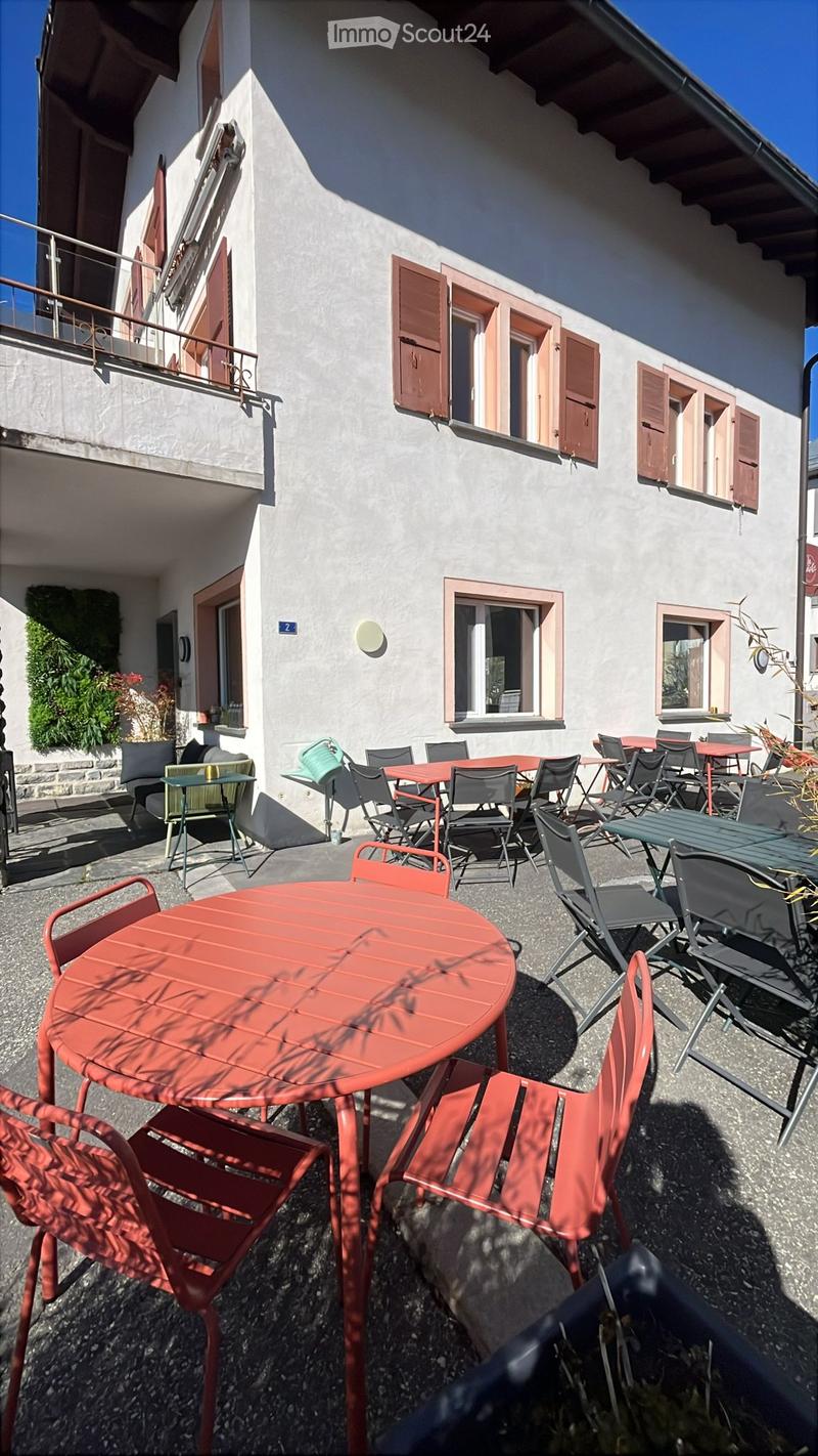 Outdoor seating area with red tables and chairs, located next to a building with shutters and a balcony.