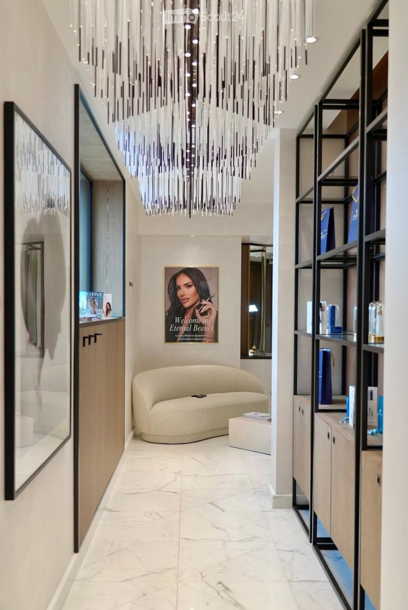 Modern, well-lit hallway with a large chandelier, a cream-colored sofa, a small table, shelves with products, and a welcome sign.