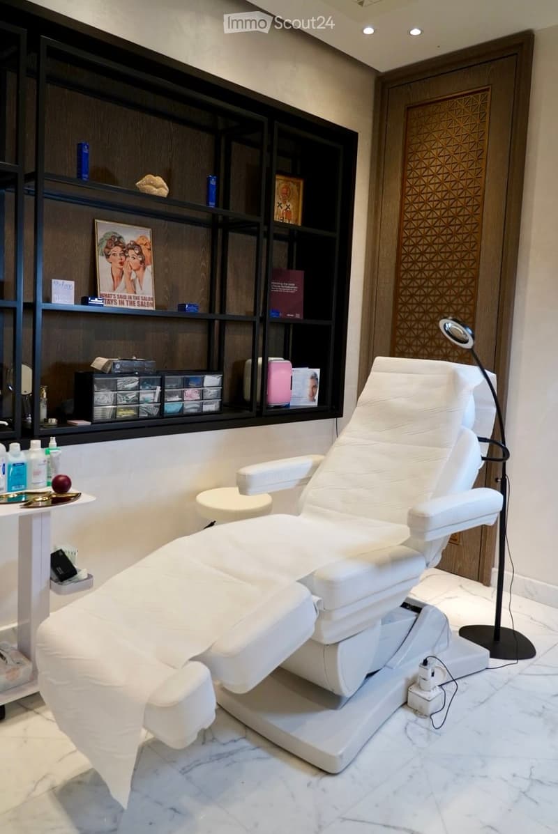A modern, well-lit salon or spa room with a white reclining chair, a small table with various items, shelves with books and products, and a decorative mirror on the wall.