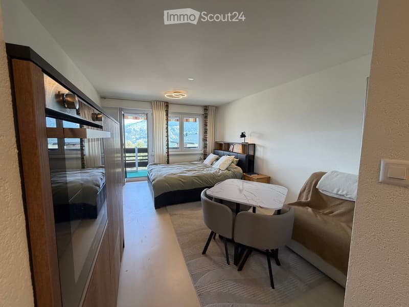 Cozy bedroom with two beds, a small table with chairs, a sofa, and a nightstand with a lamp. Large windows offer a scenic mountain view.
