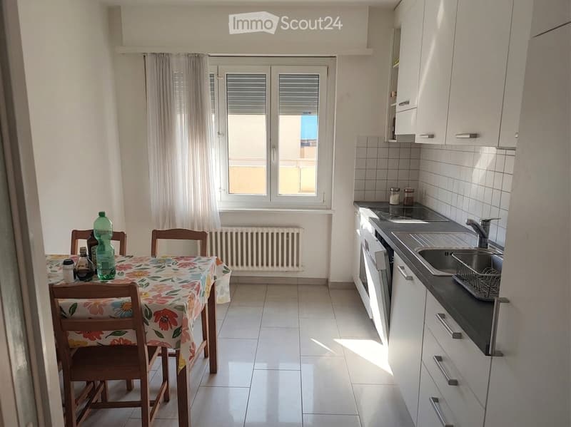 A combined kitchen and dining area with white cabinets, a black countertop, a sink, and a dining table with chairs. The room features tiled flooring and large windows with curtains.
