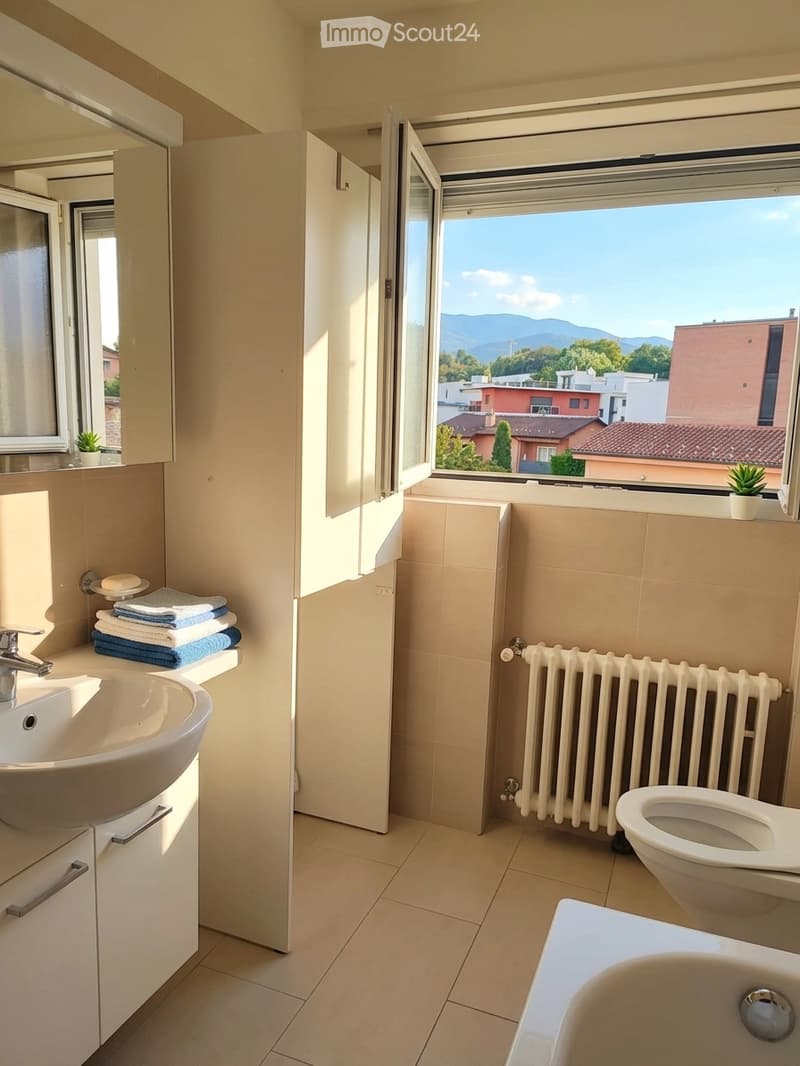 Modern bathroom with a sink, toilet, bathtub, radiator, and a large window offering a view of the city and mountains.