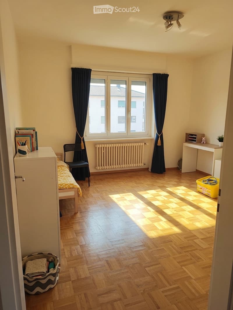Bright bedroom with wooden flooring, large window with dark curtains, bed with yellow bedding, chair, desk, radiator, and small storage items.