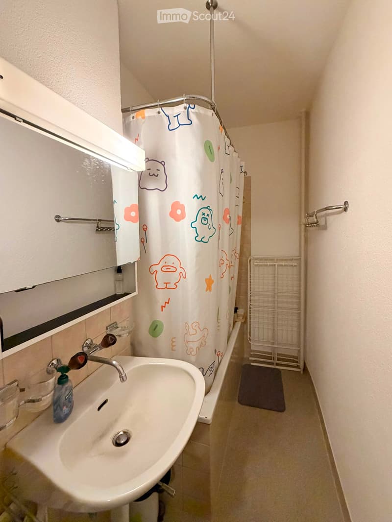 A small bathroom with a white sink, mirror, and colorful shower curtain featuring cartoon characters. A towel rack and a small shelf are visible.