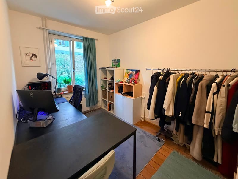 A well-lit room with a desk, computer monitor, chair, lamp, shelf, clothing rack, and a window with a curtain and plant on the windowsill.