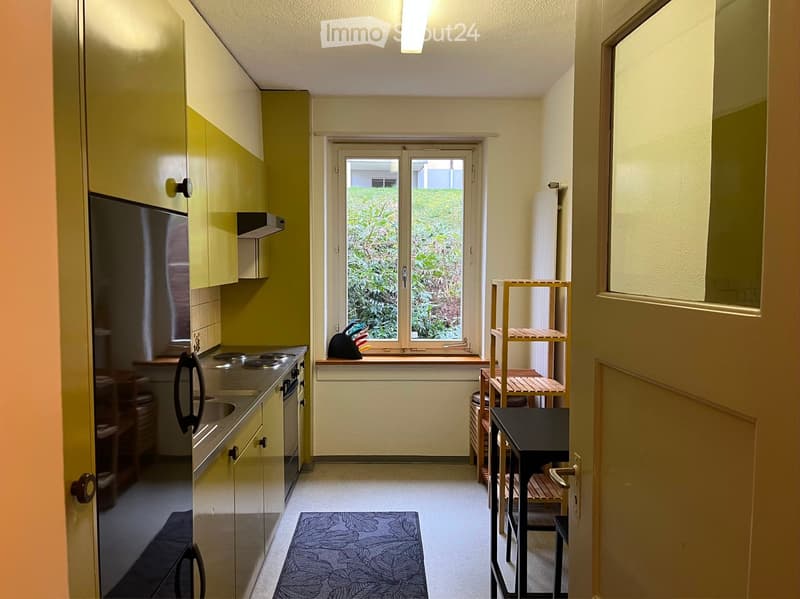 A small, modern kitchen area with light green cabinets, a stove, refrigerator, and a small dining area with a table and chairs. A window offers a view of greenery outside.