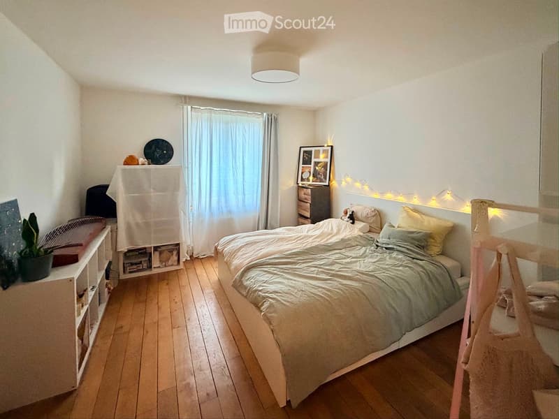 Cozy bedroom with a double bed, nightstand, potted plant, mirror, picture frame, storage unit, and a curtain covering a window. The room features wooden flooring and a slanted ceiling.