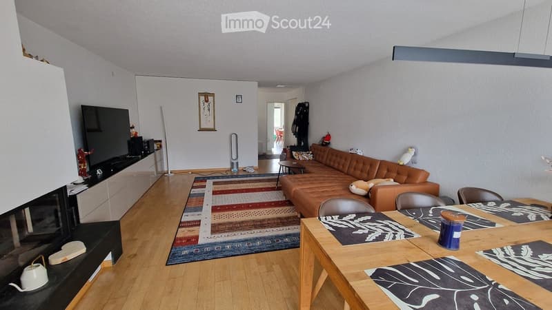 A modern living room with a brown leather sofa, wooden dining table, chairs, a television, and a floor lamp. The room features a colorful rug and a sleek black entertainment unit.