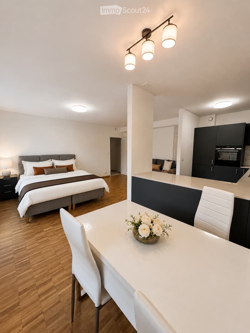 Modern, open-concept room with a bed, sofa, dining table, and kitchen area. Features wooden flooring, white walls, and contemporary lighting.