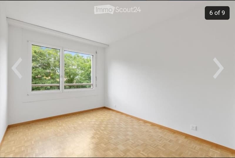 Empty room with white walls, wooden flooring, and large windows showing greenery outside.