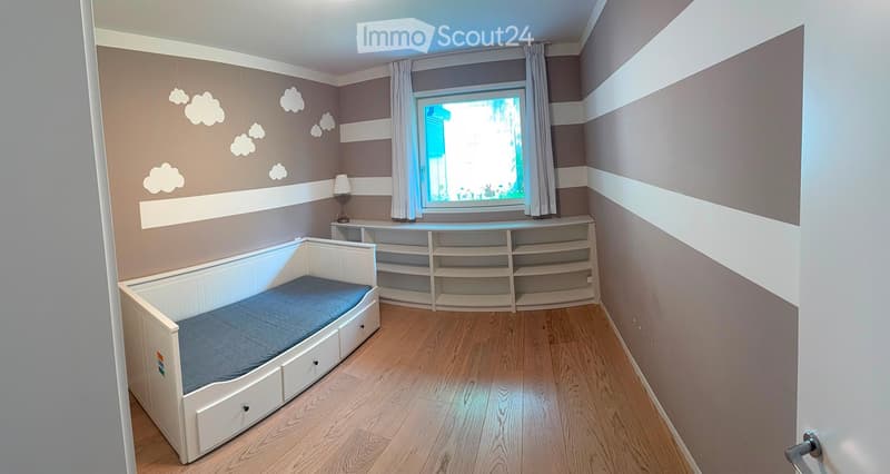 Cozy bedroom with a single bed, built-in shelving unit, and a small lamp on the shelf. The walls are painted in soft tones with cloud decals, and the floor is wooden.