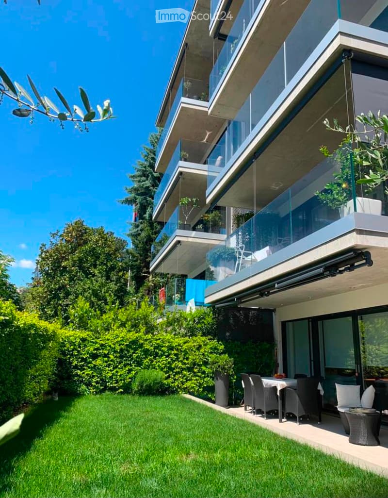 Modern multi-story building with large glass balconies, surrounded by a well-maintained garden with green lawn and shrubs. Outdoor seating area includes a table, chairs, and a sofa.