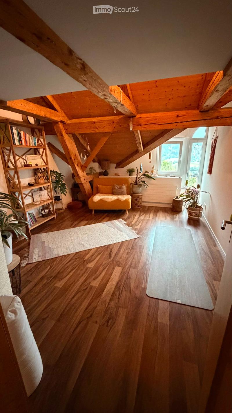 Bright, wooden-floored room with exposed wooden beams, a yellow sofa, a shelf with books and decor, a small table, a yoga mat, and several potted plants.