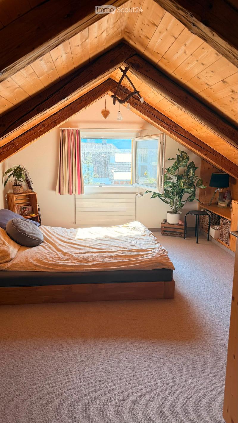 Cozy attic bedroom with wooden slanted ceilings, large windows, a double bed, nightstand, lamp, potted plants, and a small table.