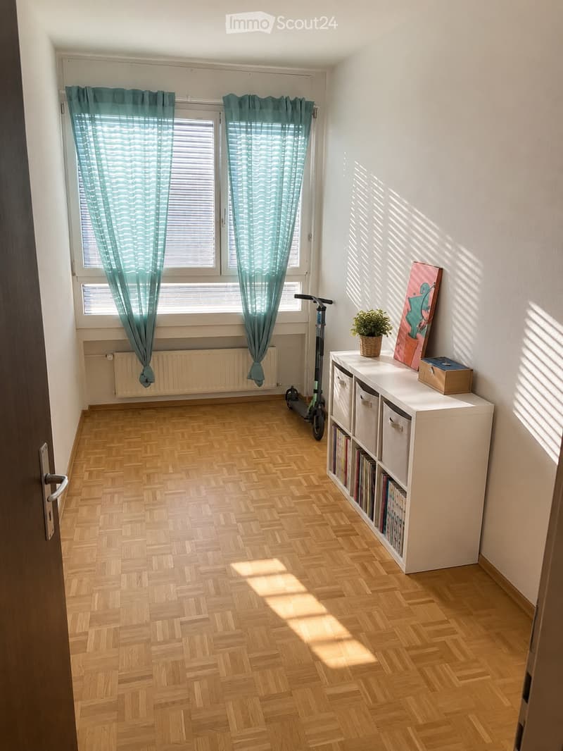 A well-lit room with wooden herringbone flooring, large windows with teal curtains, a white shelf with books and storage boxes, a potted plant, and a scooter near the window.