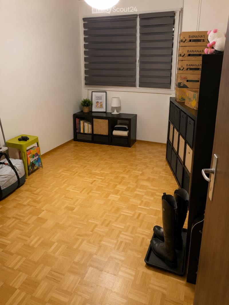 A small, well-lit room with wooden flooring, a black bookshelf, a small green stool, a lamp, a picture frame, storage boxes, and a pair of boots on the floor.