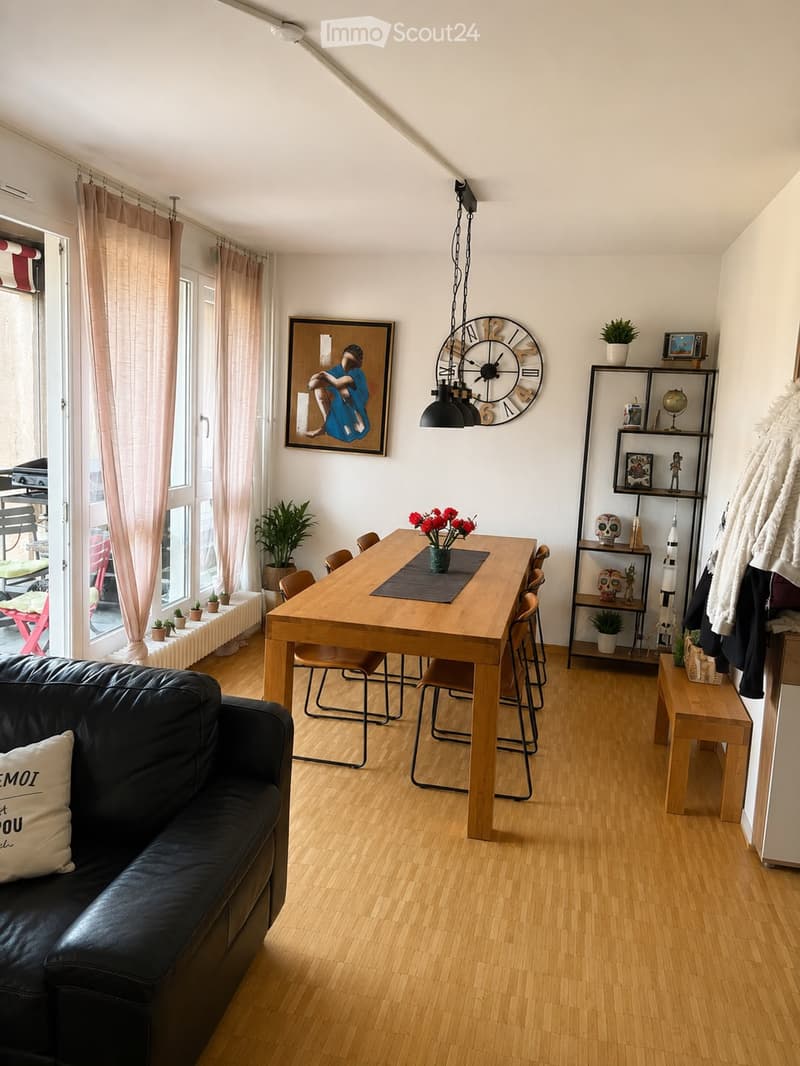 A modern, open-concept living and dining area with wooden flooring, a black leather sofa, a wooden dining table with chairs, a large wall clock, and decorative shelves with plants and artwork.