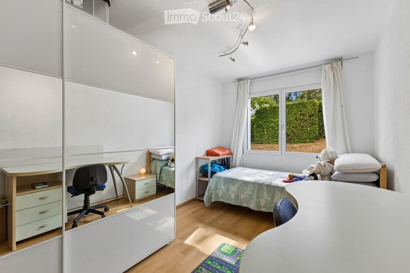 A well-lit bedroom with a bed, desk, chair, wardrobe, and nightstand. The room features wooden flooring and large windows with white curtains, allowing natural light to fill the space.