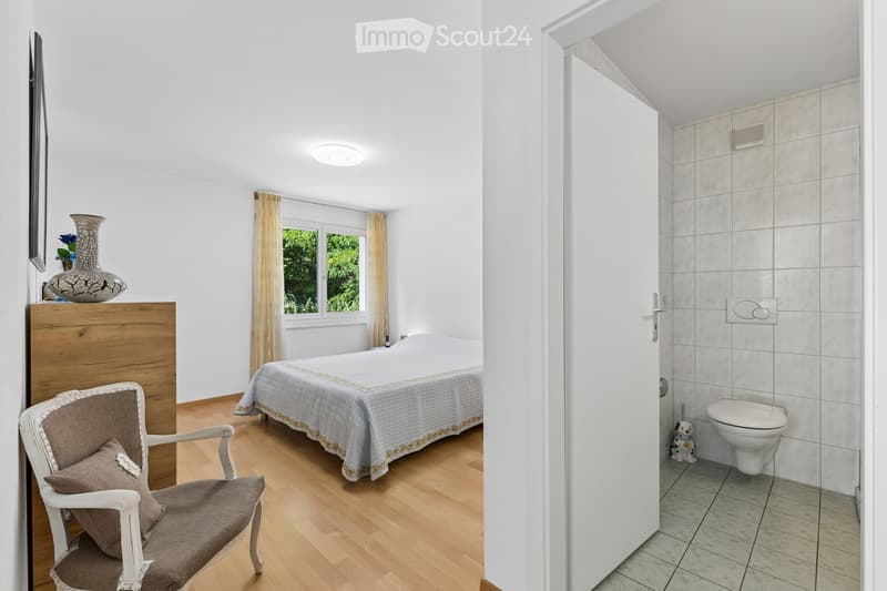 Cozy bedroom with a wooden bed, a white armchair with a brown cushion, a decorative vase on a wooden nightstand, and a small bathroom with a toilet visible through an open door.