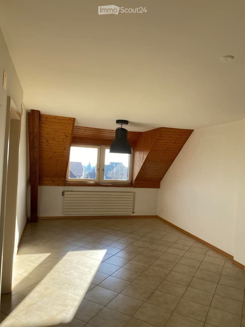 Empty room with wooden slanted ceiling, tiled floor, white walls, window with wooden frame, radiator, and a single hanging light fixture.