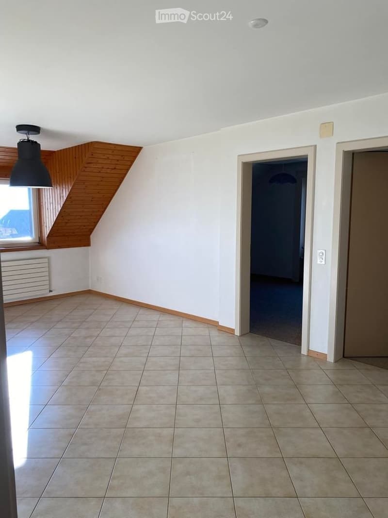 Empty room with tiled floor, white walls, and wooden slanted ceiling. Features include a window with wooden frame, radiator, and two doors leading to other spaces.