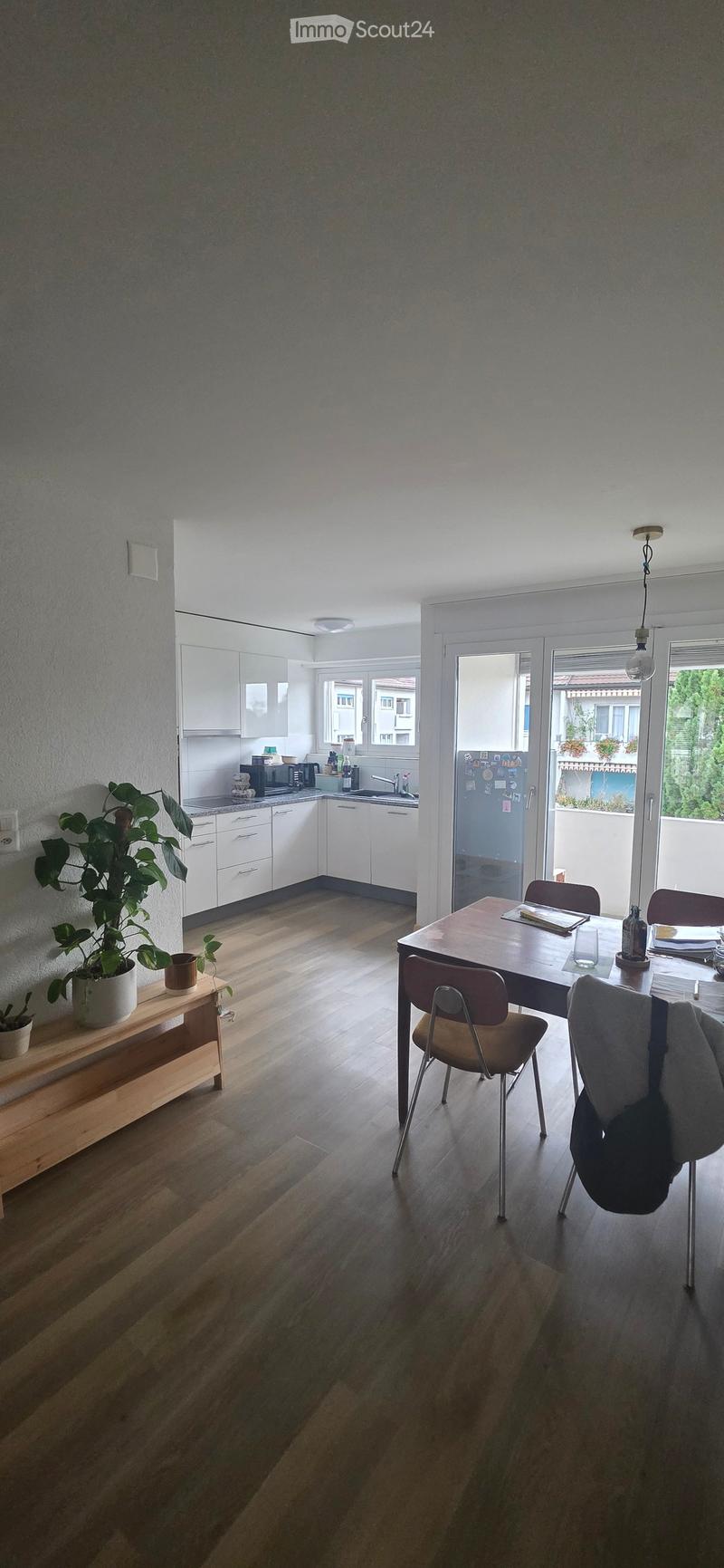 A modern kitchen and dining area with white cabinets, a wooden dining table, chairs, and a potted plant on a sideboard. The kitchen features a sink, microwave, and refrigerator, with large windows and a sliding glass door leading to a balcony.