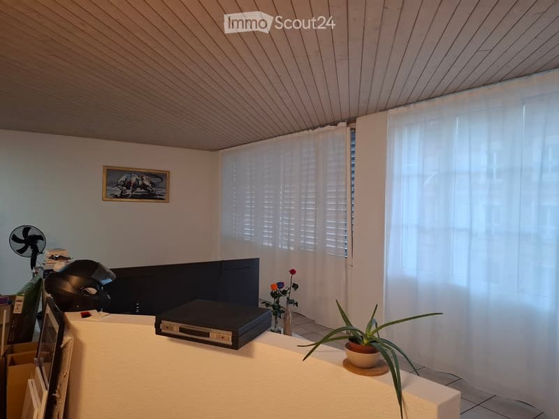 A furnished room with a slanted wooden ceiling, white walls, and large windows covered by white curtains. Visible objects include a table with a potted plant, a fan, a picture frame, a vase with flowers, and a screen.