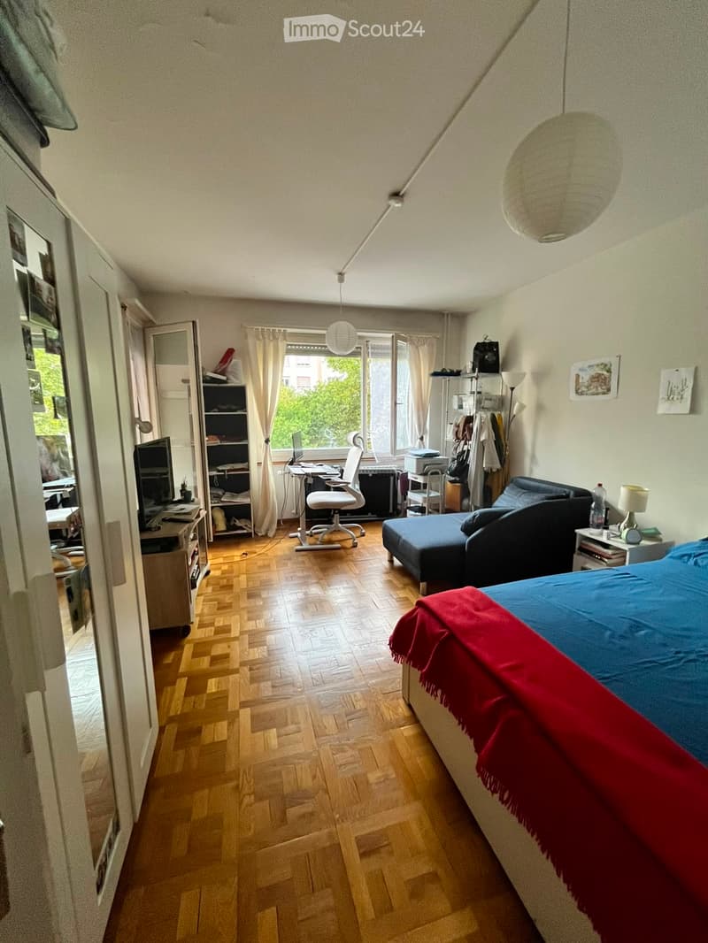 A furnished room with a bed, sofa, chair, table, laptop, shelf, and lamp. The room features wooden flooring and large windows with curtains.