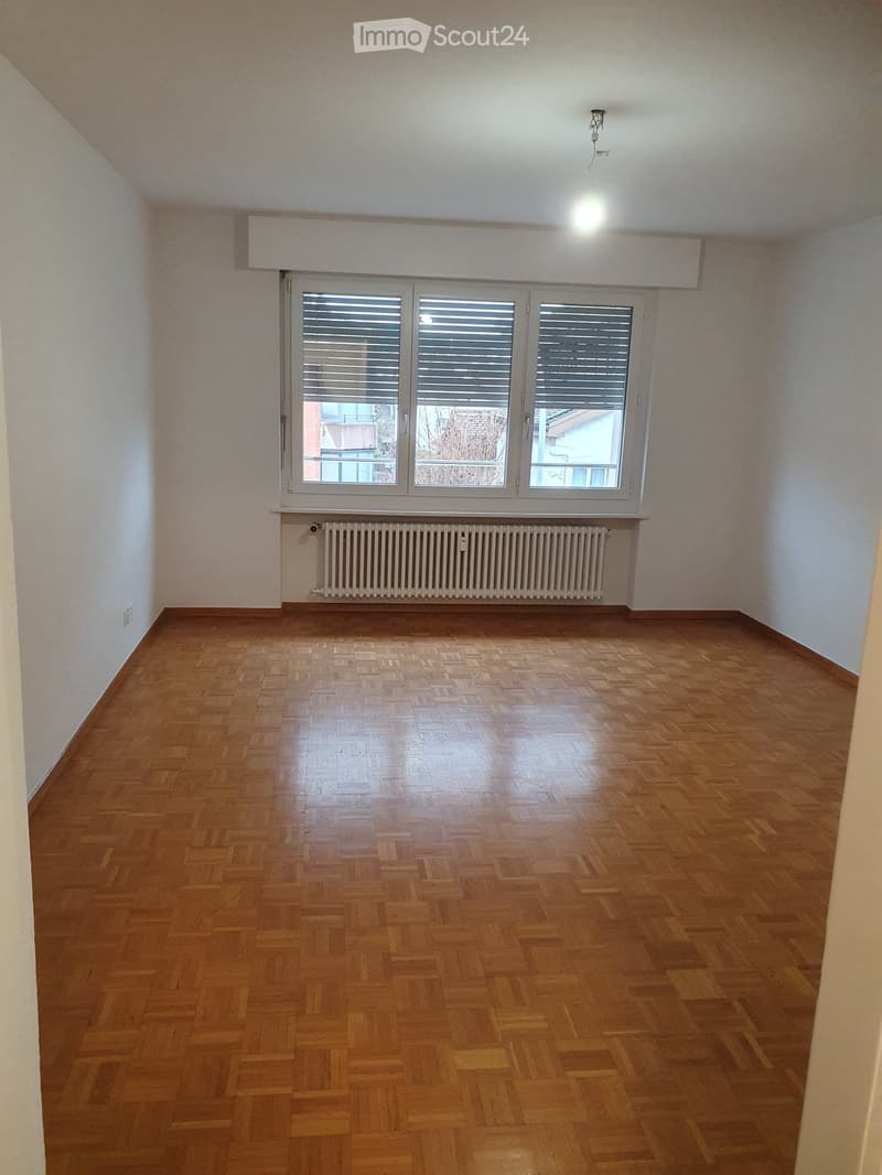 Empty room with wooden parquet flooring, white walls, large window with blinds, radiator, and ceiling light fixture.