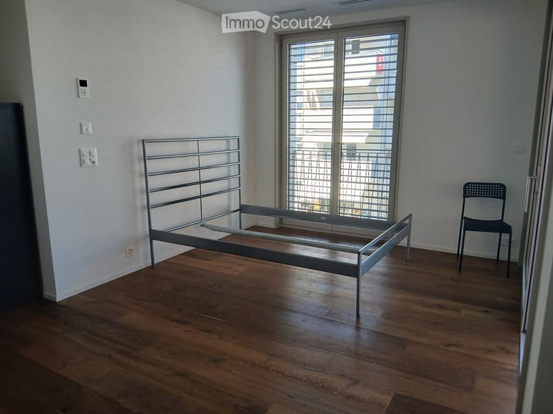 Empty bedroom with wooden flooring, a metal bed frame, and a single chair near the window.
