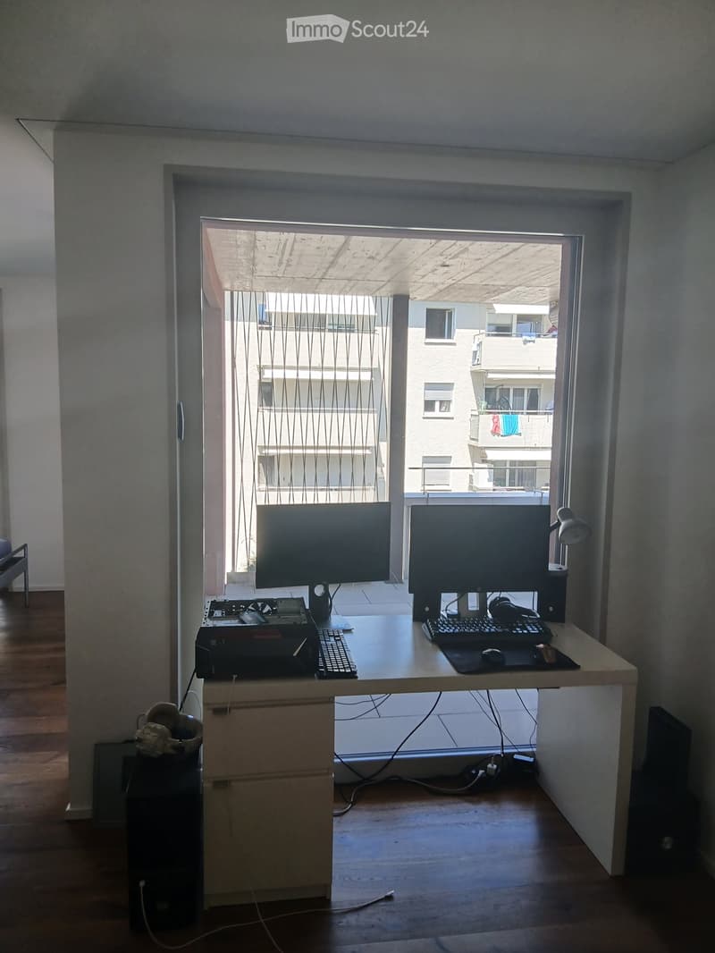 A modern, minimalist workspace with a large desk, dual monitors, keyboard, mouse, printer, and headphones. The room features wooden flooring and large windows with sheer curtains, offering a view of the building exterior.