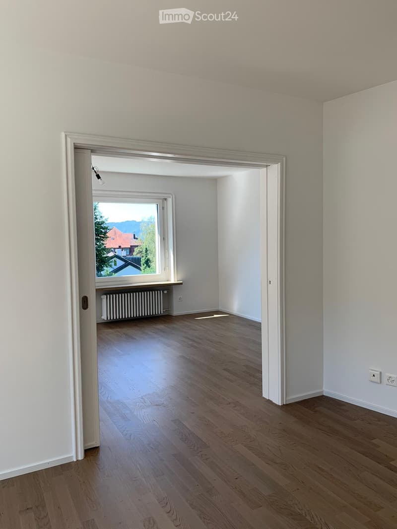 Empty room with light wooden flooring and white walls, featuring a doorway leading to another room with a window and radiator visible.