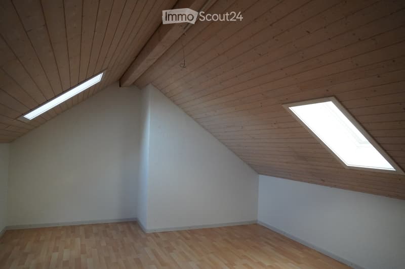 Empty room with wooden floor and slanted wooden ceiling, featuring two skylight windows and white walls.