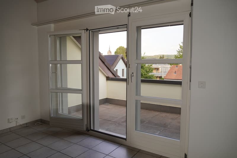 Empty room with tiled floor and large sliding glass doors opening to a balcony area. The balcony has a tiled surface and overlooks neighboring buildings and greenery.
