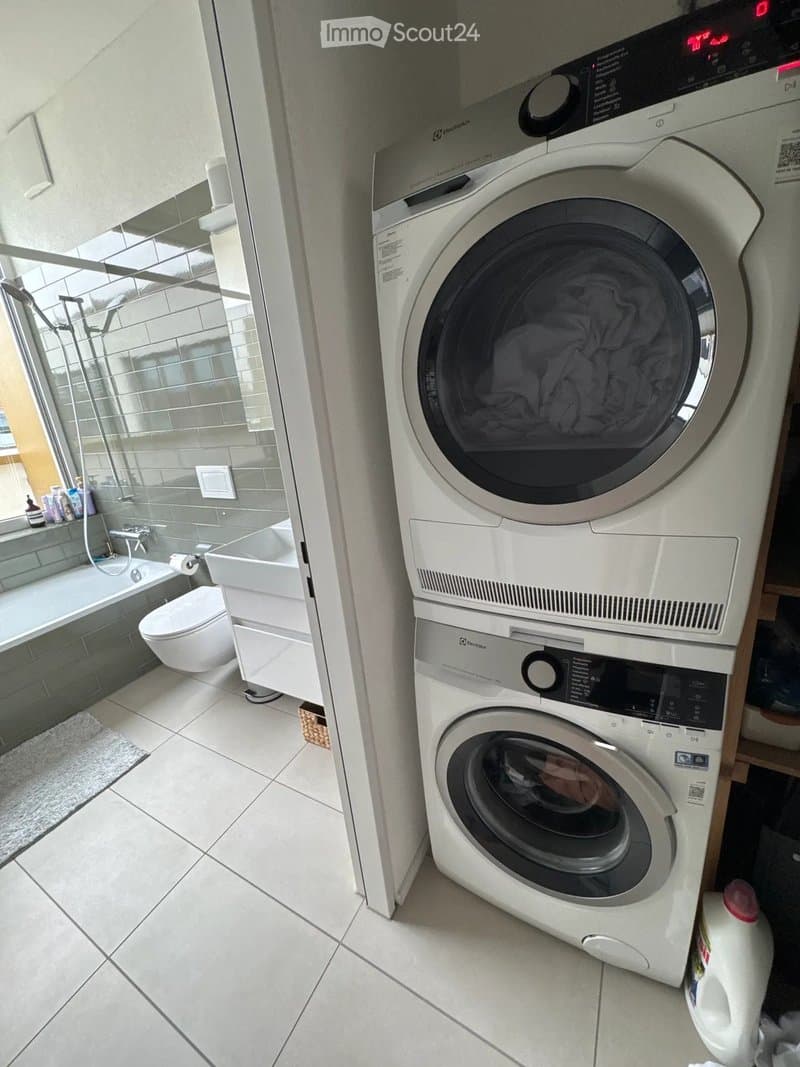 Modern bathroom with a white tiled floor, a toilet, a bathtub, a shower area, and a stacked washer and dryer unit.