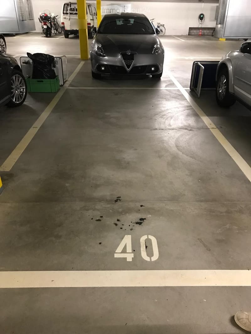 Underground parking lot with several parked cars, motorcycles, and bicycles. The floor is marked with yellow lines, and there are yellow pillars and a green crate visible.