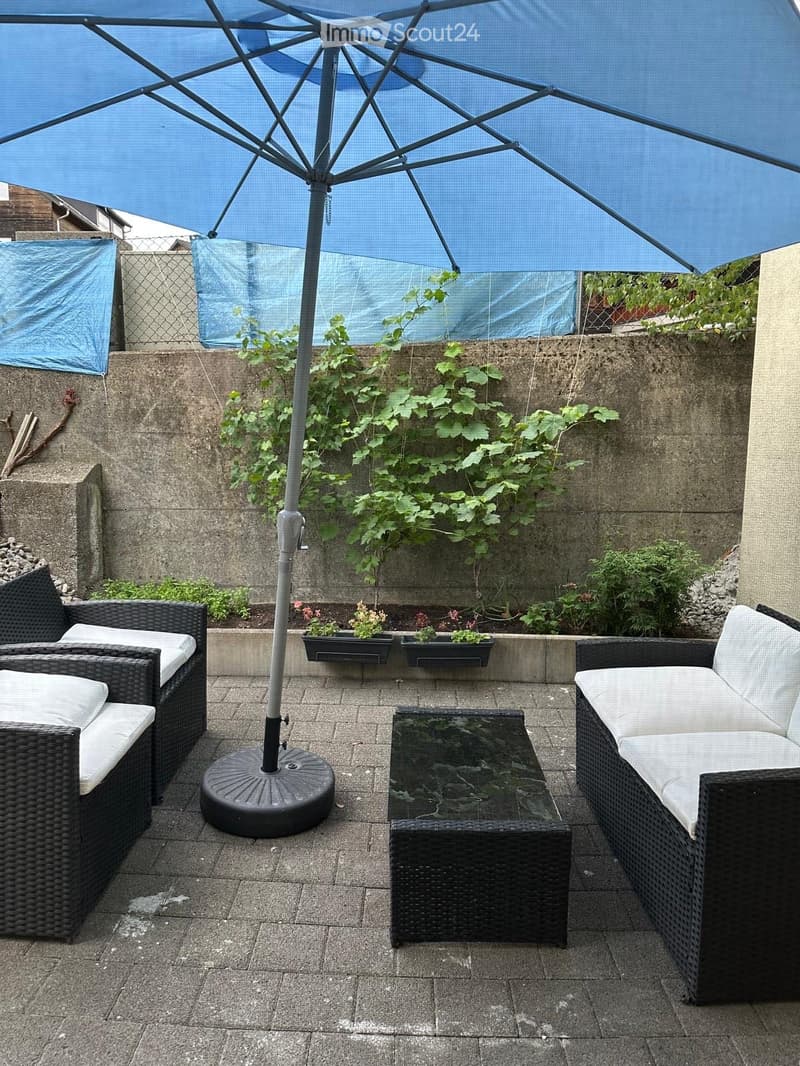 A small outdoor patio with a large umbrella, two wicker sofas, a glass coffee table, and potted plants.