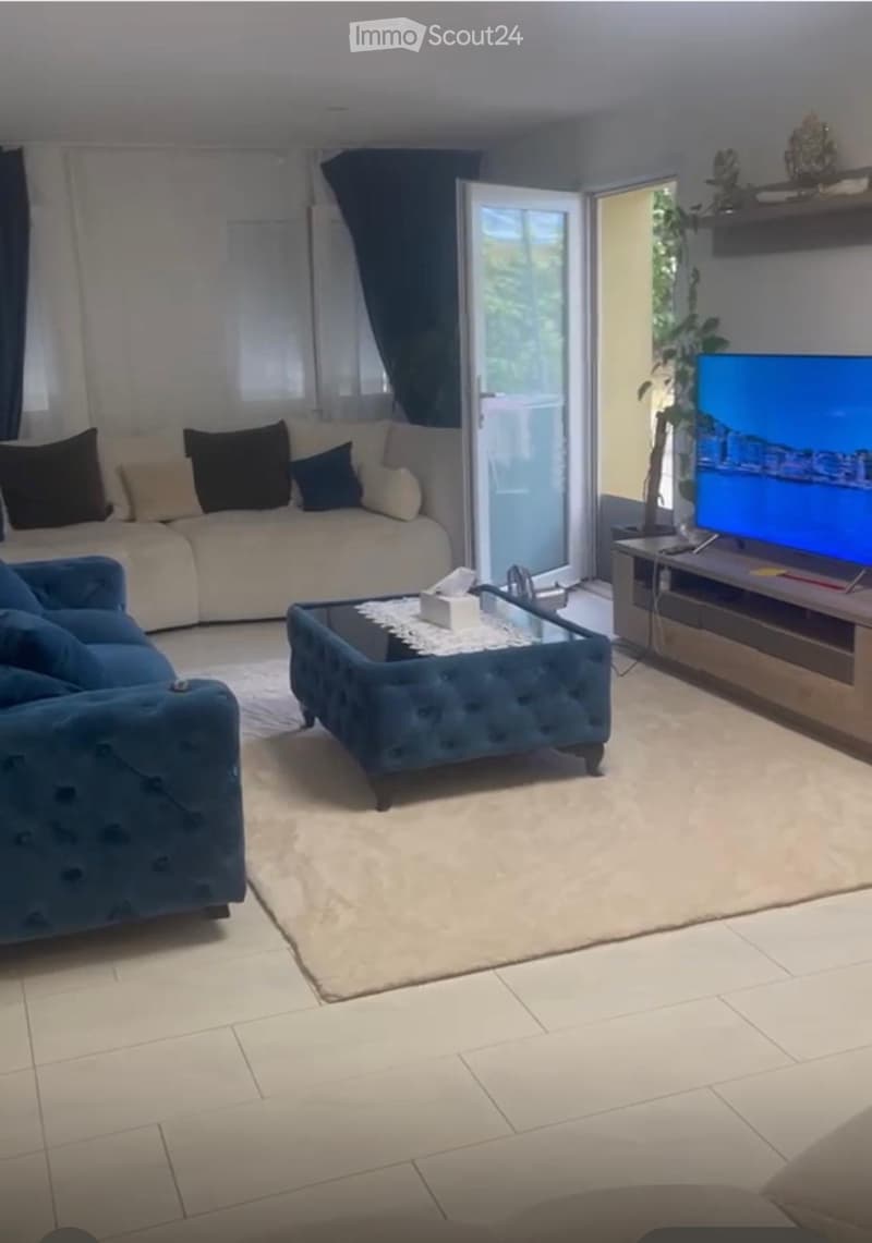 Modern living room with a beige sofa, blue armchair, glass coffee table, flat-screen TV, and large windows leading to a balcony.