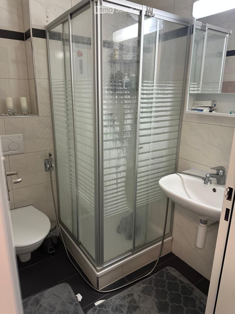 Modern bathroom with a glass-enclosed shower, toilet, sink, and mirror. Includes a small shelf with candles and a grey bath mat on the floor.