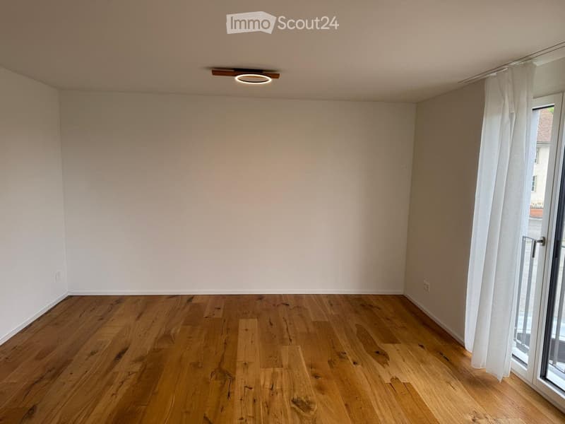 Empty room with wooden flooring, white walls, and a large window with white curtains.