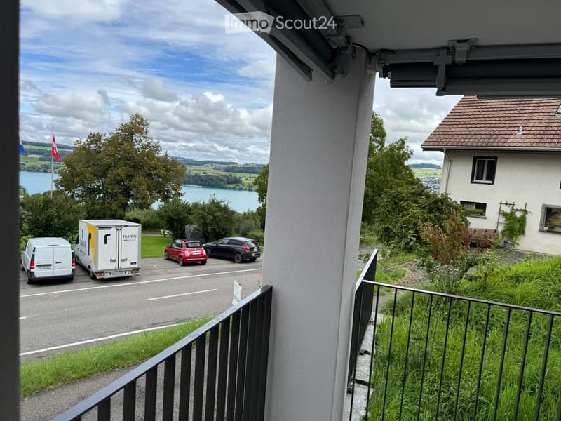 Balcony overlooking a street with parked cars, a moving van, and a scenic view of a lake and surrounding landscape.