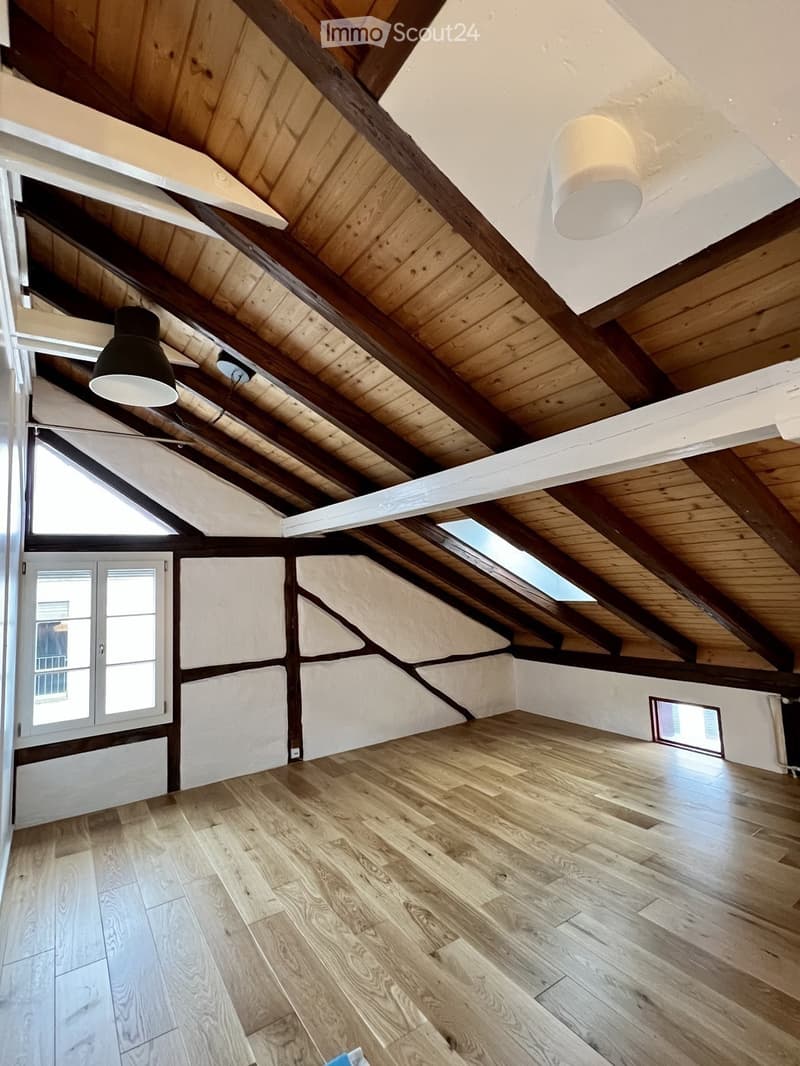 An empty attic space with wooden beams, exposed wooden ceiling, wooden floor, and skylights. The walls are white with a textured finish, and there are two windows on the left side.