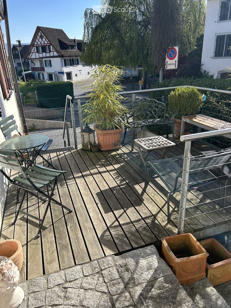 Balcony with wooden decking, metal railing, outdoor furniture including a table, chairs, bench, lantern, and potted plants.