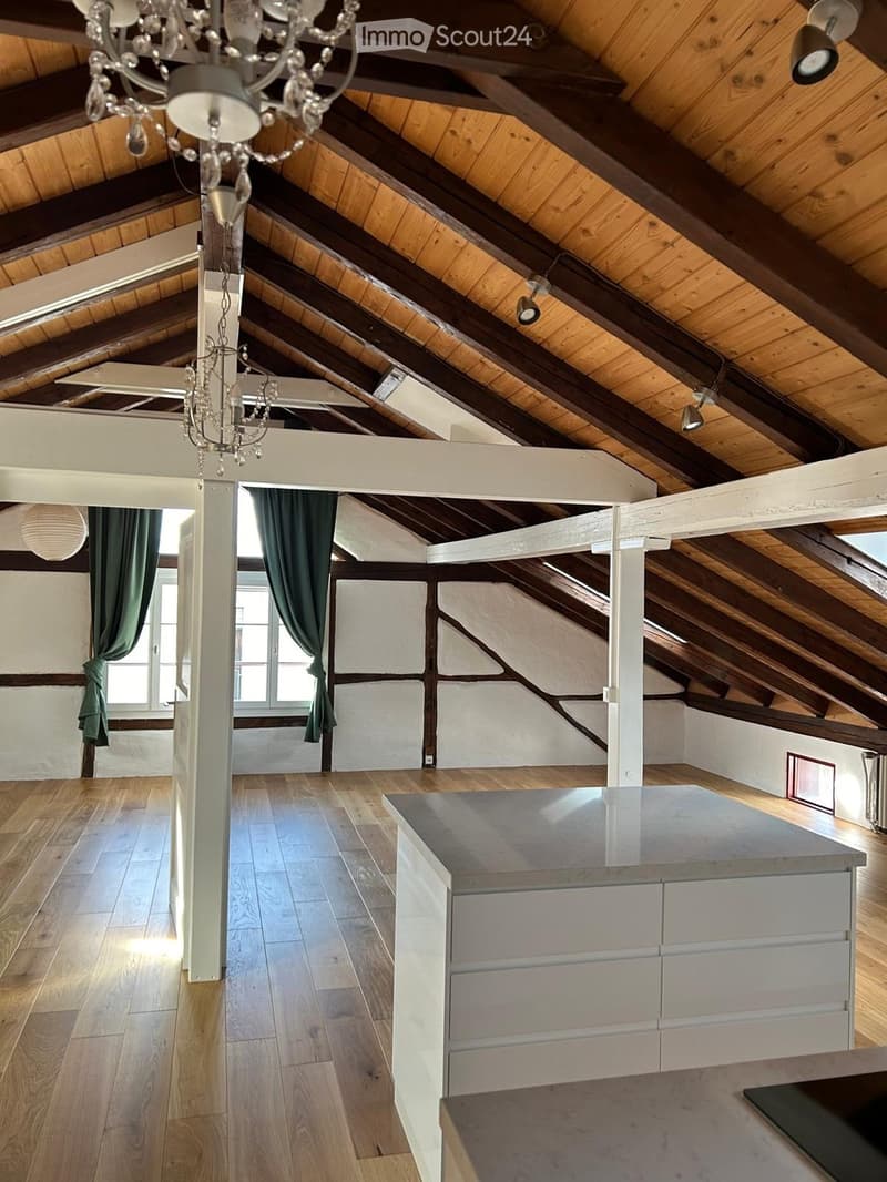 Open-plan attic space with exposed wooden beams, wooden flooring, and a modern kitchen island. Green curtains on windows, and a chandelier hanging from the ceiling.