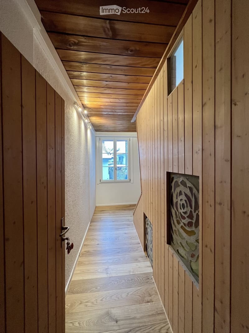 Wooden-paneled hallway with a slanted wooden ceiling, recessed lighting, and a window at the end. Features a decorative stained-glass panel on one wall and a door with a keyhole on the left.