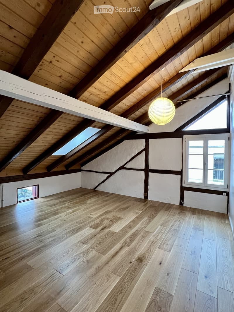 Empty attic space with wooden floors, exposed wooden beams, skylights, and large windows, featuring white walls and a hanging light fixture.