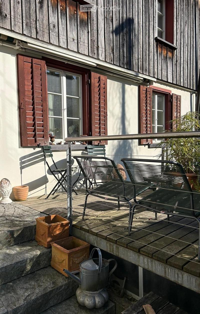 Balcony with a metal railing, wooden deck, and outdoor furniture including a table, chairs, and a bench. Potted plants and a watering can are placed on the steps leading up to the balcony.