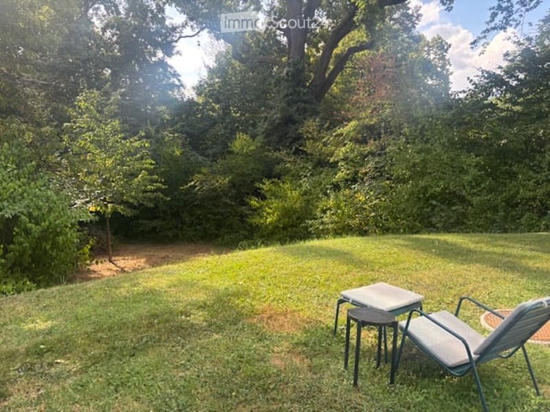 A well-maintained garden with a small seating area featuring a table and a chair, surrounded by lush greenery and trees.