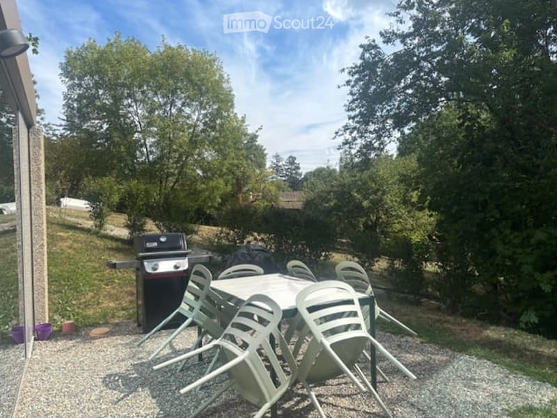 Outdoor patio area with a round table, several chairs, and a grill, surrounded by trees and greenery.
